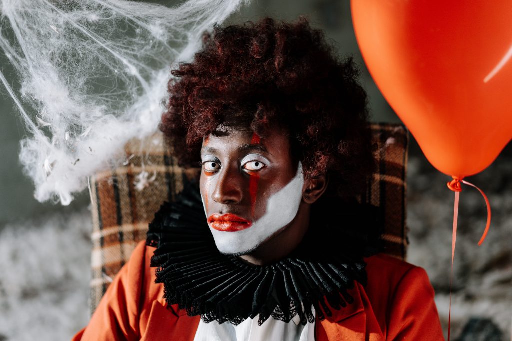 person in clown makeup seated in a tweed-covered armchair side eyes the viewer