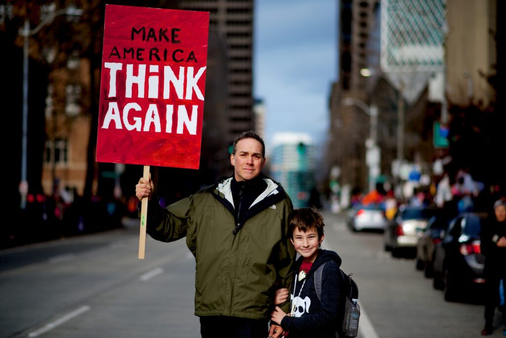 child stands by person holding red and white protest sign