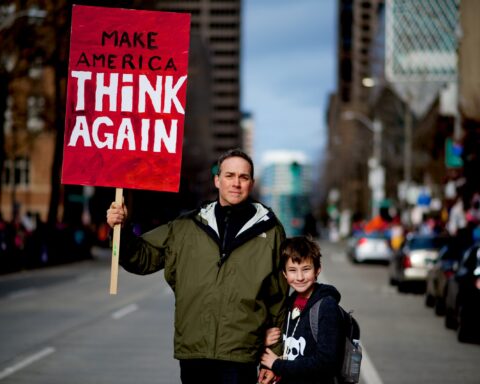 child stands by person holding red and white protest sign